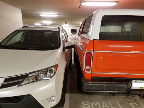 1978 Ford Bronco - An orange 1978 Ford Bronco is parked closely beside a white SUV in a parking garage.