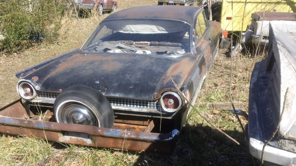 Rusty black 1960 Ford Starliner with a spare tire mounted on the front, seen from a low front angle