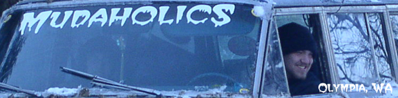 Person wearing a beanie smiles from inside a vehicle with a "MUDAHOLICS" sticker on the windshield. "OLYMPIA, WA" text is visible
