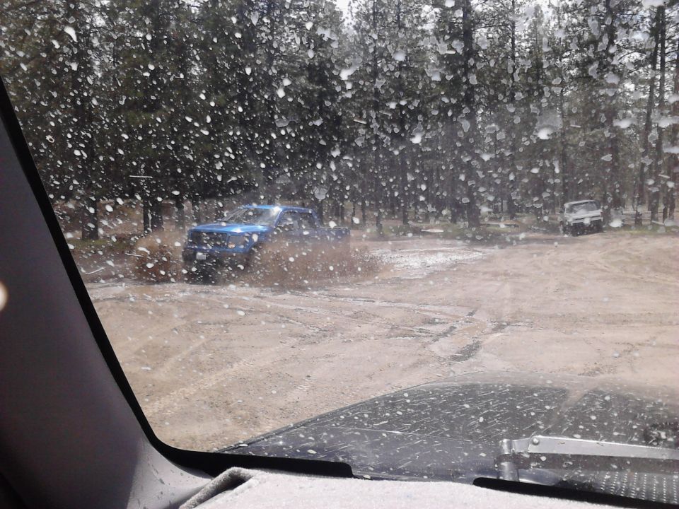 Blue pickup truck splashing through a muddy trail with another vehicle in the background