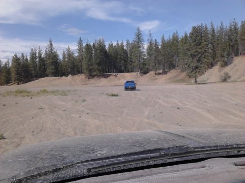 1995 Ford Bronco - A lone blue pickup truck sits in a dusty, open area surrounded by trees under a partly cloudy sky.