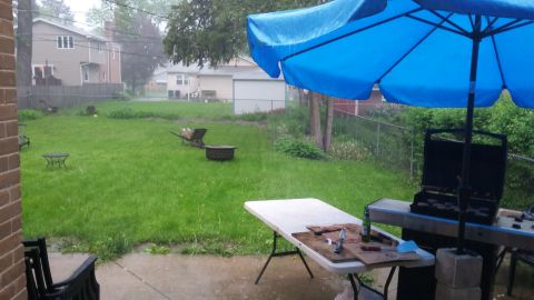 Backyard scene with a grill under a blue umbrella, a table with food, and lawn chairs