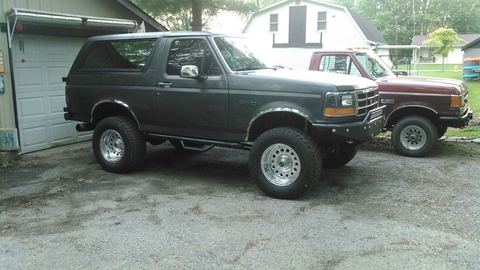 Lifted gray Ford Bronco on polished wheels in a gravel driveway beside a red F-150