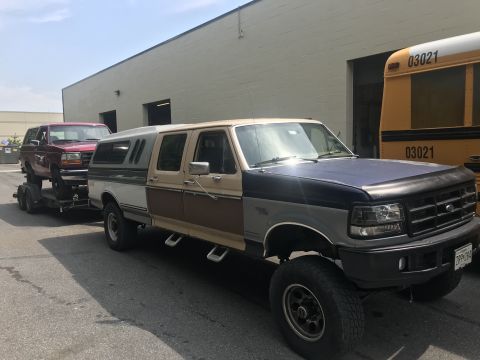 1994 Ford Bronco - A lifted 1994 Ford F-250 truck tows a red Ford Bronco on a trailer, with a yellow school bus visible in the background.