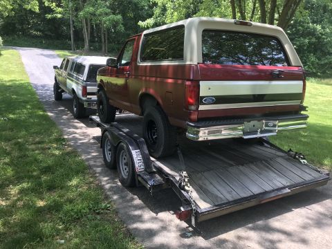 1994 Ford Bronco - A red and tan Ford Bronco is being towed on a trailer, with another truck following behind it on a paved road.