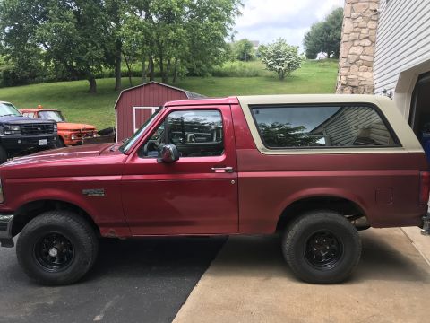1994 Ford Bronco - Red Ford Bronco with tan top parked next to a garage. Other vehicles are visible in the background.
