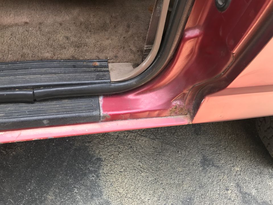 Close-up of a red vehicle's door sill with some rust and worn trim