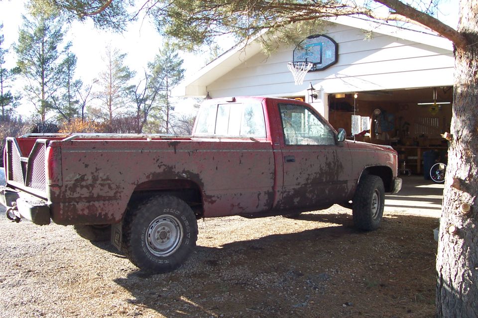 Muddy dark red single-cab 4x4 pickup parked rear-side view near open garage