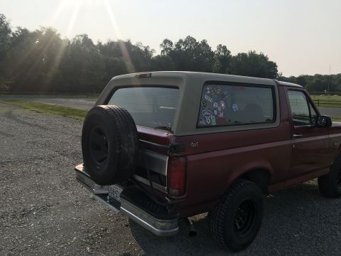 1994 Ford Bronco - A maroon Ford Bronco is parked on a gravel surface with trees in the background.