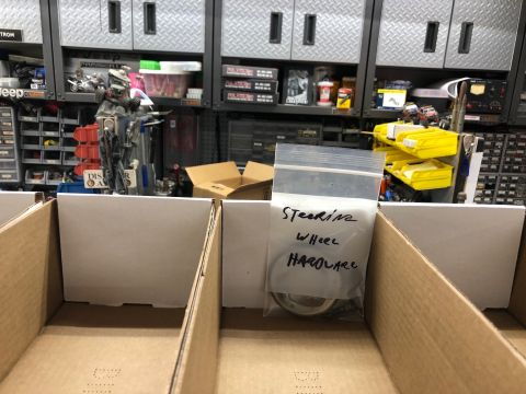 1992 Jeep Wrangler - A plastic bag containing steering wheel hardware is placed in a cardboard box, with shelves of parts in the background.