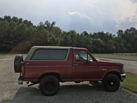 1994 Ford Bronco - Side profile of a red 1994 Ford Bronco with a tan top and spare tire mounted externally.
