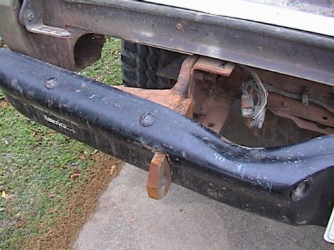 1978 Ford Bronco - Low-angle rear detail of a truck's black rear bumper, showing rusty steel mounting brackets bolted to the frame rails and a receiver tube b…