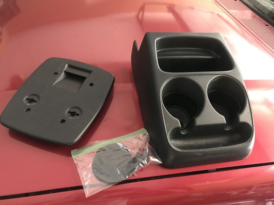 Black plastic automotive console with cup holders and a separate mounting plate on a red surface.