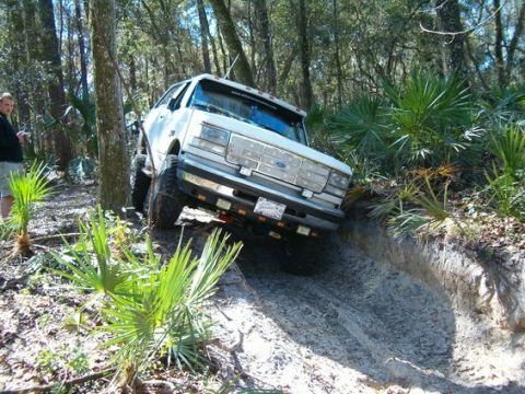 1995 Ford Bronco - Lifted white Ford Bronco flexing at a steep angle as it drops over a sand ledge on a wooded Florida trail lined with saw palmetto. The 3/4…