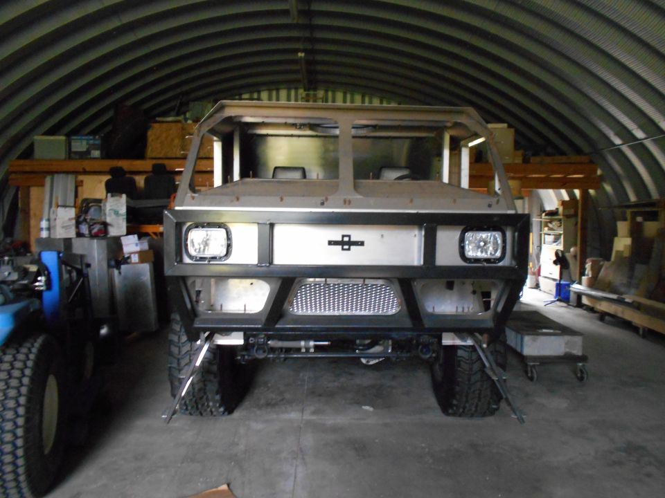 Front view of a custom vehicle build in a workshop, showing bumper, headlights, and cab frame.