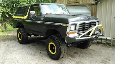 1978 Ford Bronco - A lifted black 1978 Ford Bronco with yellow accents on the wheels and trim is parked on a gravel surface in front of a wooden building.