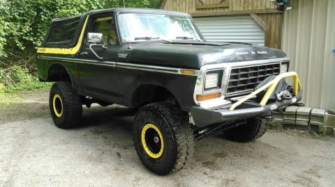 1978 Ford Bronco - A lifted, dark green 1978 Ford Bronco with yellow accents sits parked on a gravel driveway in front of a wooden building. The Bronco featur…