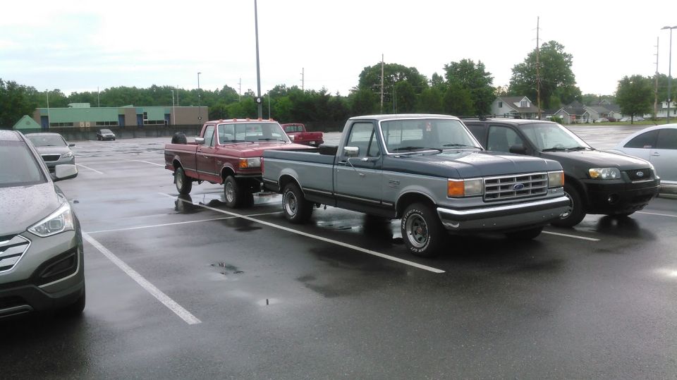 Two pickup trucks parked in a wet lot. Foreground is a gray and blue Ford F-150, background is a red Ford truck.