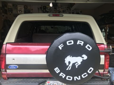 1994 Ford Bronco - The rear of a red and tan Ford Bronco is shown, featuring a spare tire cover with the Ford Bronco logo and a horse silhouette. The vehicle…