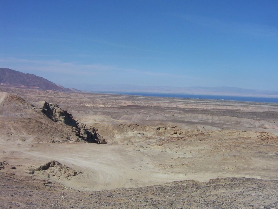 Desert landscape with mountains, dry terrain, and a distant body of water under a clear blue sky. Watermark reads SUPERMOTORS