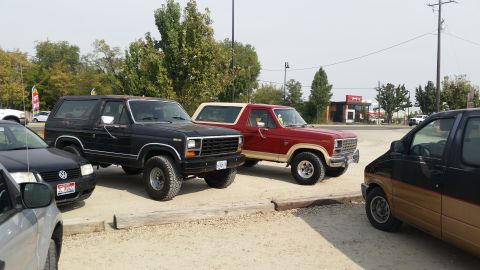 1985 Ford Bronco - A black 1980s Ford Bronco is parked next to a red and tan Ford Bronco. A black Volkswagen Jetta is parked in front of the black Bronco.