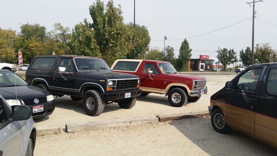 Two 1980s Ford Broncos and a black Volkswagen Jetta parked on a dirt lot.