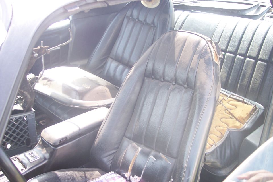 Interior of a classic car showing worn black leather bucket seats and a center console with a large object in the middle.