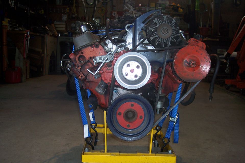 Red V8 engine on a yellow stand with serpentine belt, alternator, and power steering pump.