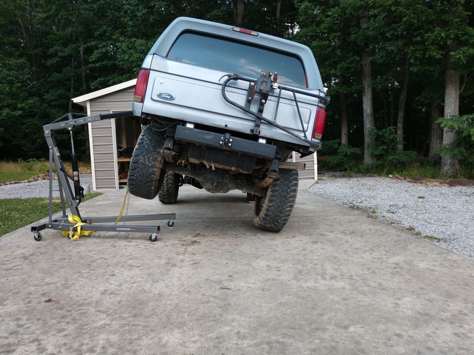 Silver Ford Bronco lifted by engine hoist on concrete driveway.