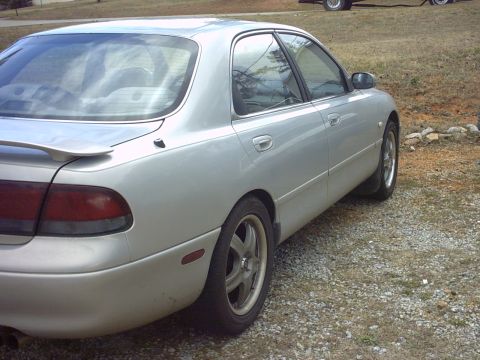 1995 Mazda 626 - A silver sedan is viewed from the rear quarter angle, parked outdoors on a gravel surface.