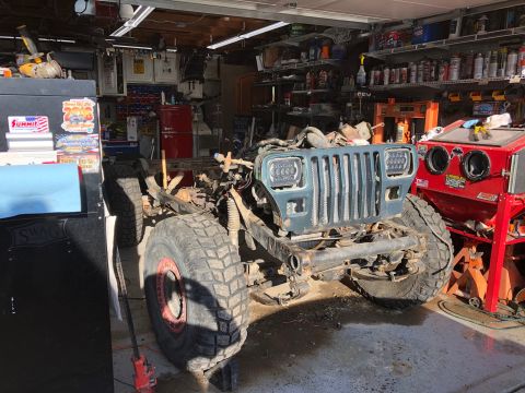 1992 Jeep Wrangler - A disassembled Jeep Wrangler chassis with large off-road tires is parked in a cluttered garage. The front grille and headlights are visible…