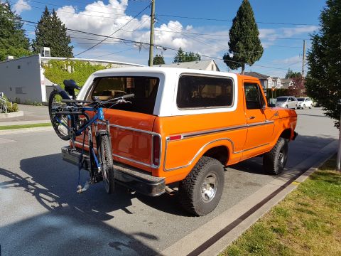 1978 Ford Bronco - A bright orange 1978 Ford Bronco with a white roof is parked on a residential street, carrying a blue bicycle on a rear-mounted rack. The v…