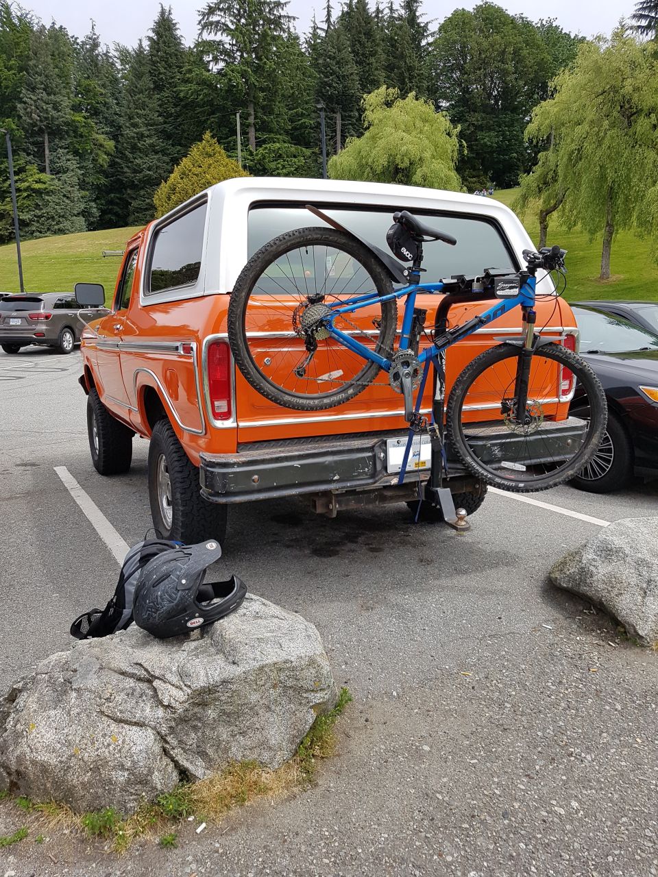 Orange 1978 Ford Bronco with a blue mountain bike on a rear rack, parked next to a black car. A helmet is on a rock.