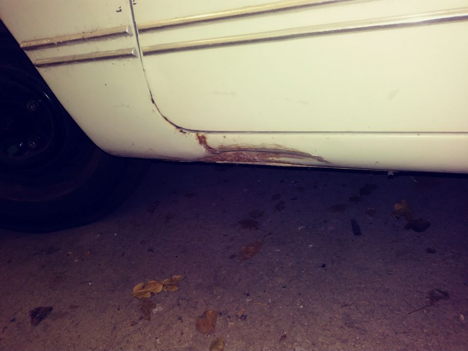 White vehicle rocker panel with rust damage, tire, and ground.