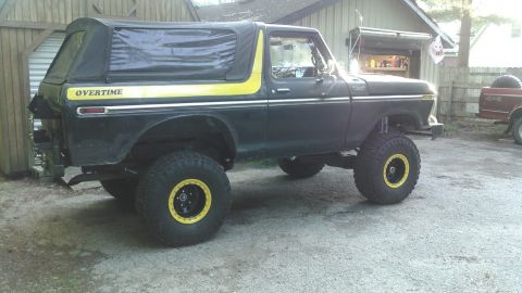 1978 Ford Bronco - A lifted black 1978 Ford Bronco with a black soft top and yellow "OVERTIME" decal on the side is parked on a gravel driveway next to a wood…