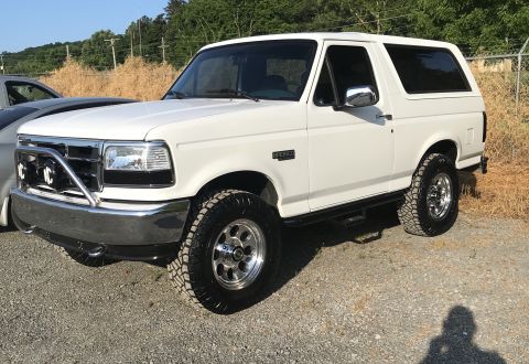 1995 Ford Bronco - A white 1990s Ford Bronco is parked on a gravel lot with dry grass and trees in the background. The SUV has a chrome grille guard with KC l…