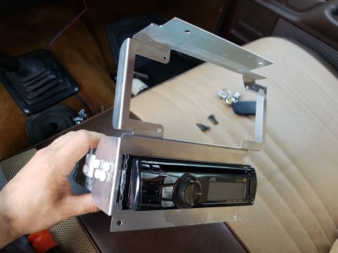 1978 Ford Bronco - A hand holds a JVC car stereo mounted in a custom aluminum bracket, positioned in front of a vehicle's tan interior seats. The bracket appe…