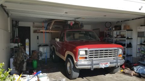 1985 Ford Bronco - A red 1980s pickup truck is parked inside a garage with a Bronco or similar vehicle on top of it. The truck is facing forward and is parked…