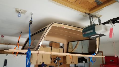1985 Ford Bronco - A truck cap is suspended from the ceiling of a garage by straps and a wooden plank. A garage door opener unit is mounted to the ceiling abo…