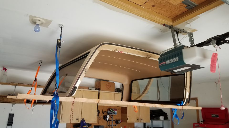 Truck cap suspended from garage ceiling by straps and wood plank. Garage door opener visible.