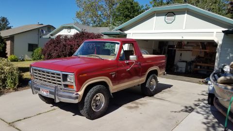 1985 Ford Bronco - A red 1980s Ford Bronco is parked in a driveway in front of a garage. The Bronco has tan trim and off-road tires.