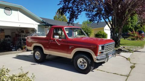 1985 Ford Bronco - A red and tan 1980s Ford Bronco is parked in a driveway in front of a garage. The SUV has off-road tires and a spare tire visible in the be…