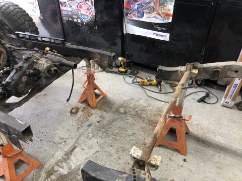 1992 Jeep Wrangler - A vehicle frame is supported by jack stands in a garage setting. A transmission and part of a tire are visible on the left side of the fram…