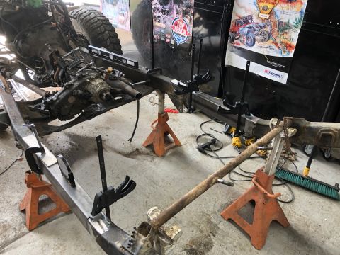1992 Jeep Wrangler - The chassis of a Jeep Wrangler is supported by jack stands in a garage. The transmission and transfer case are visible, along with parts of…