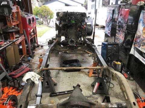 1992 Jeep Wrangler - The chassis of a Jeep Wrangler is elevated on jack stands in a garage setting, with the engine and transmission assembly still attached. A…