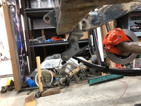 1992 Jeep Wrangler - Rear of a vehicle chassis on stands showing a leaf spring, shackle, and orange brake drum, surrounded by tools in a cluttered garage.