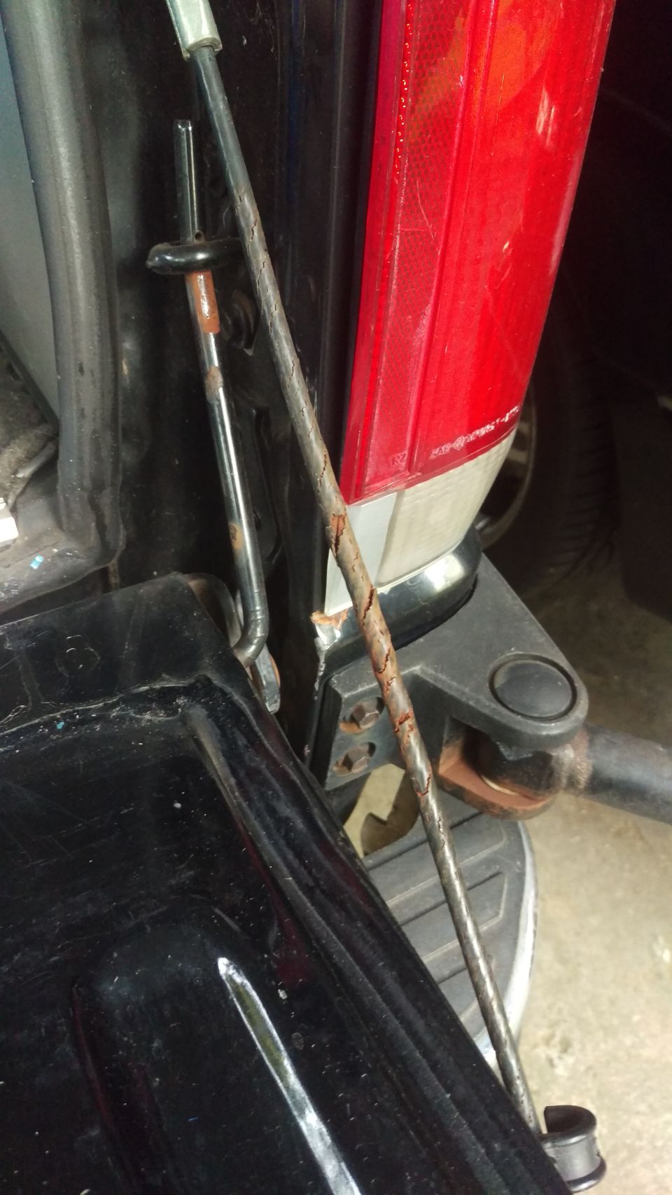 Close-up of rusted tailgate support cable and red taillight on a vehicle.