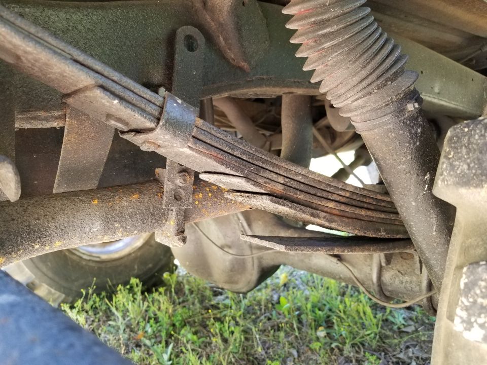 Rusty leaf spring pack with a broken leaf under a vehicle, viewed from below with grass beneath