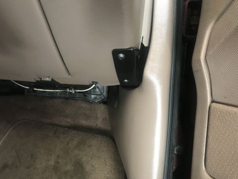 1994 Ford Bronco - A black metal bracket with two silver screws is mounted to the beige interior paneling of a vehicle, near the floor and door jamb.