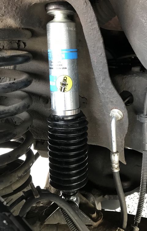 1995 Ford Bronco - A close-up view of a silver Bilstein shock absorber with a black accordion boot installed on a vehicle. The shock absorber has blue and yel…